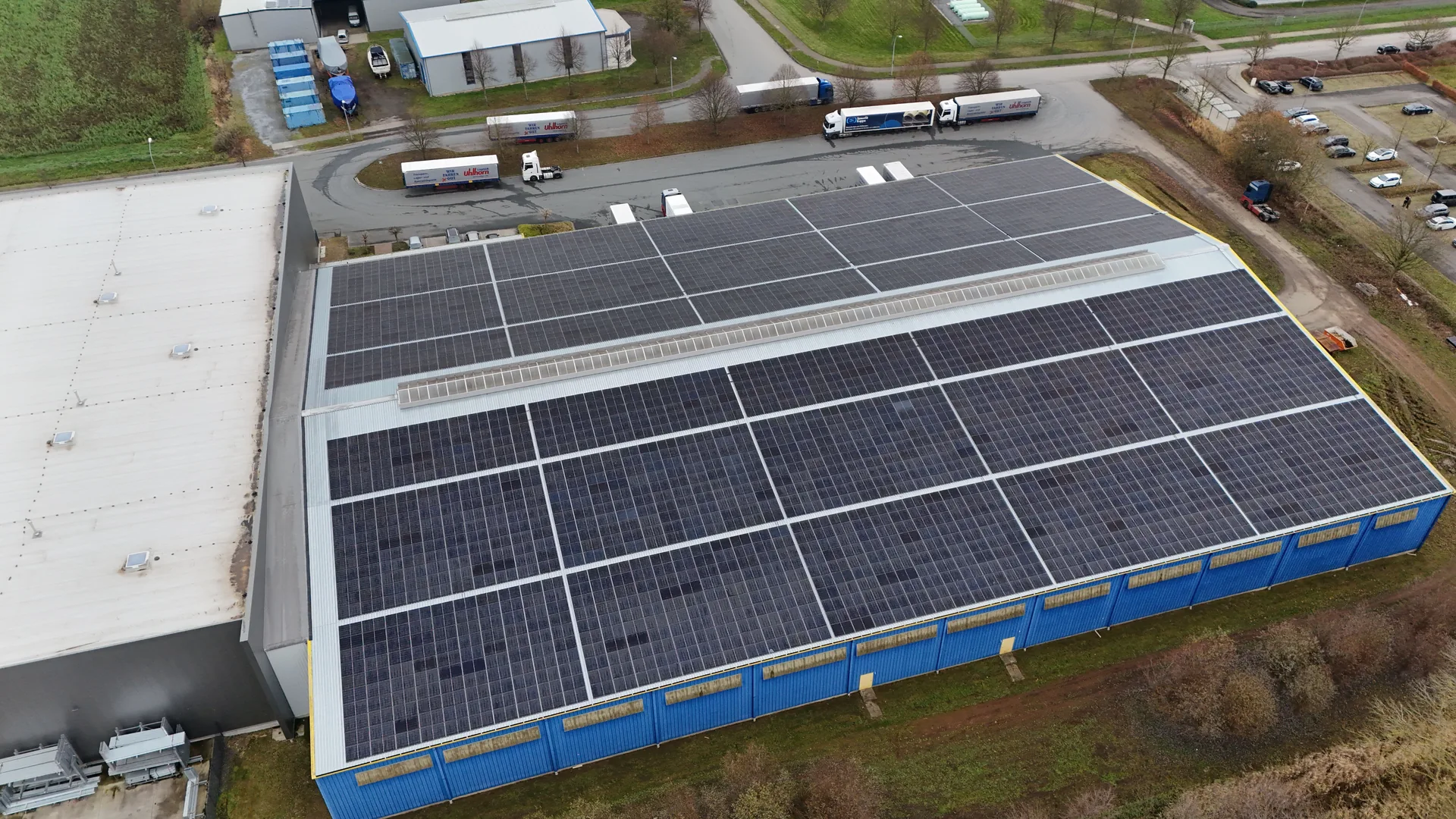 Vertikale Draufsicht auf das vollständig mit Solarmodulen belegte Hallendach, LKWs und Trailer im Hintergrund