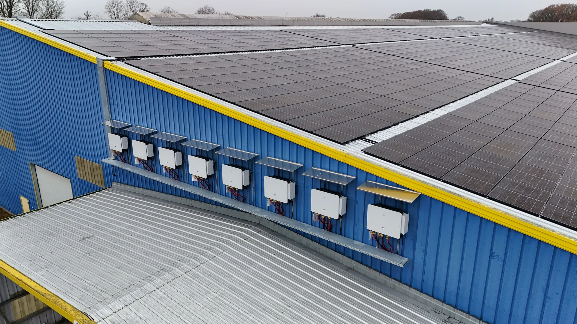 Sieben wandmontierte Wechselrichter an der blauen Hallenfassade, darüber die Modulreihen des Daches